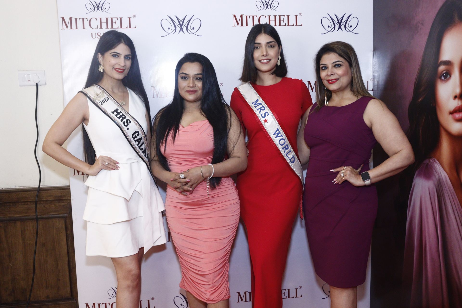 Four women posing in formal dresses at an event with Mitchell backdrop, two wearing sashes.